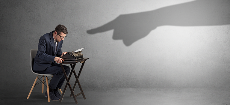 Giant shadow finger pointing at an employee who is an insider threat. Man is in a suit sitting at a desk on a chair. Grey background.