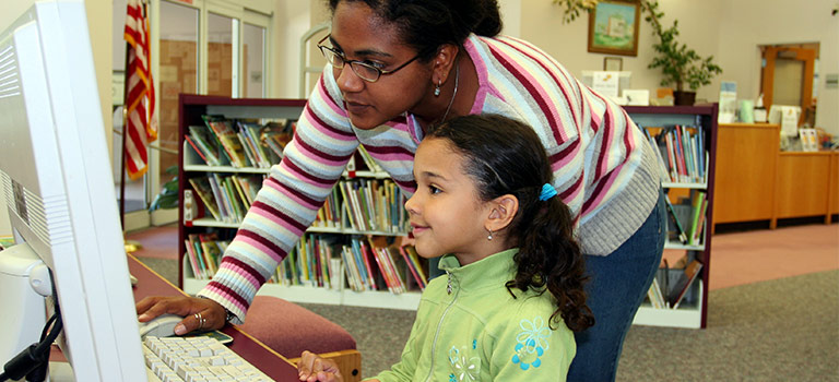 Woman assisting young girl who is typing on a computer in a library child internet safety